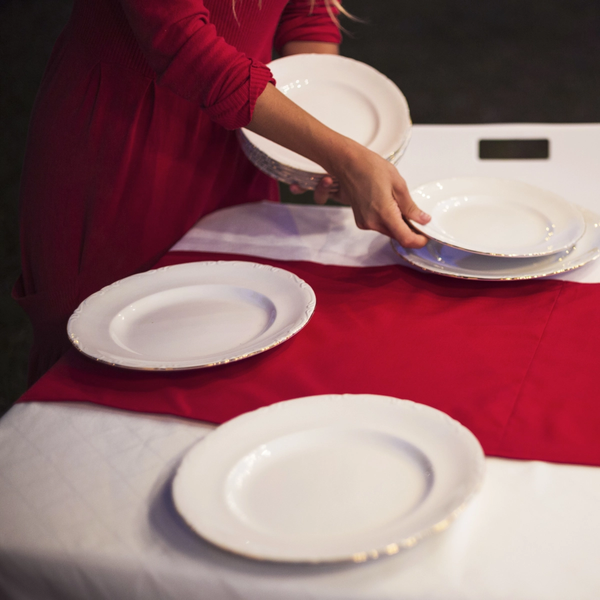 Woman setting white plates on a red and white holiday tablecloth outdoors.