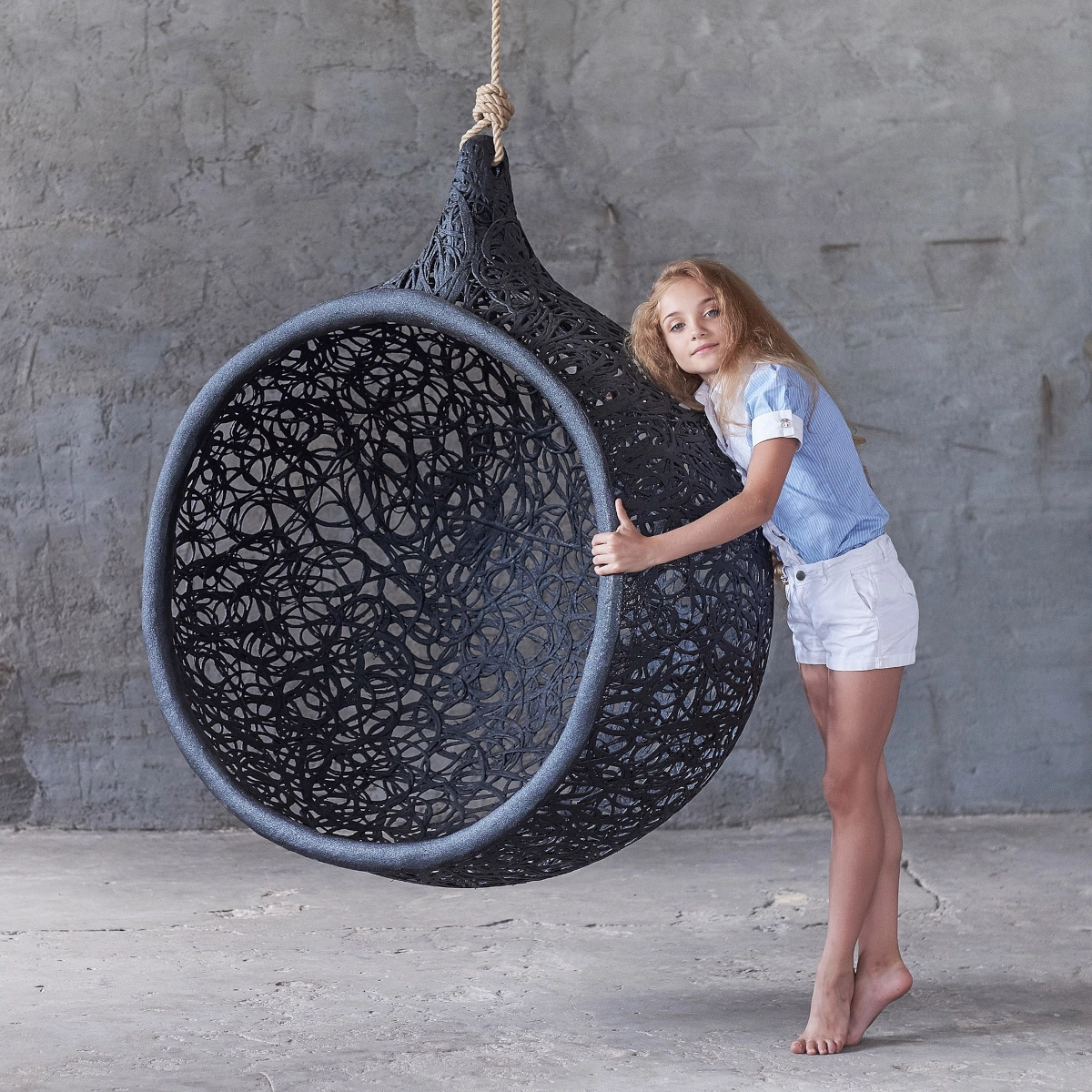 Girl hugging modern black hanging egg chair secured with rope from ceiling.