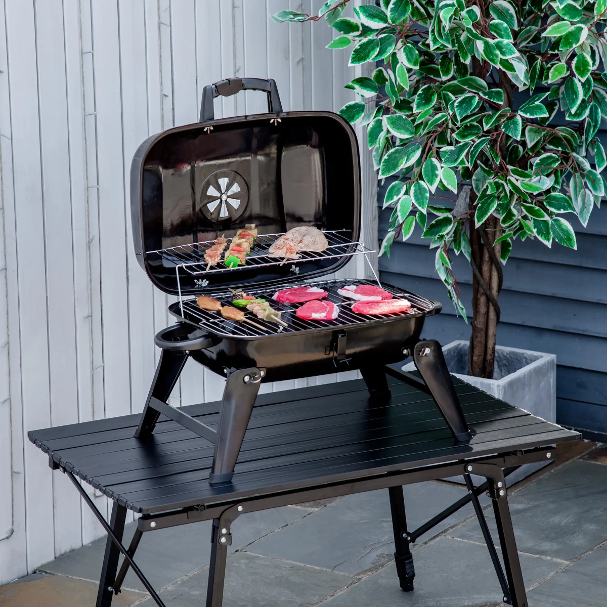 Portable tabletop charcoal grill with meat and skewers, perfect for camping and backyard BBQs.