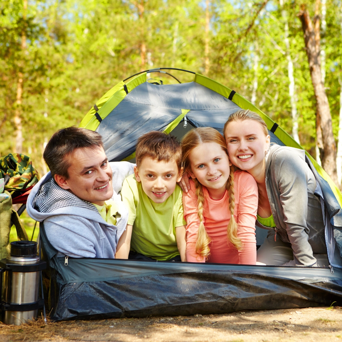 Family smiling inside green camping tent in forest with thermos and backpacks outside.