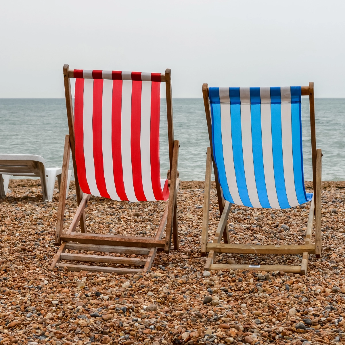 Red and blue striped wooden beach chairs facing the ocean on a pebbled shore.