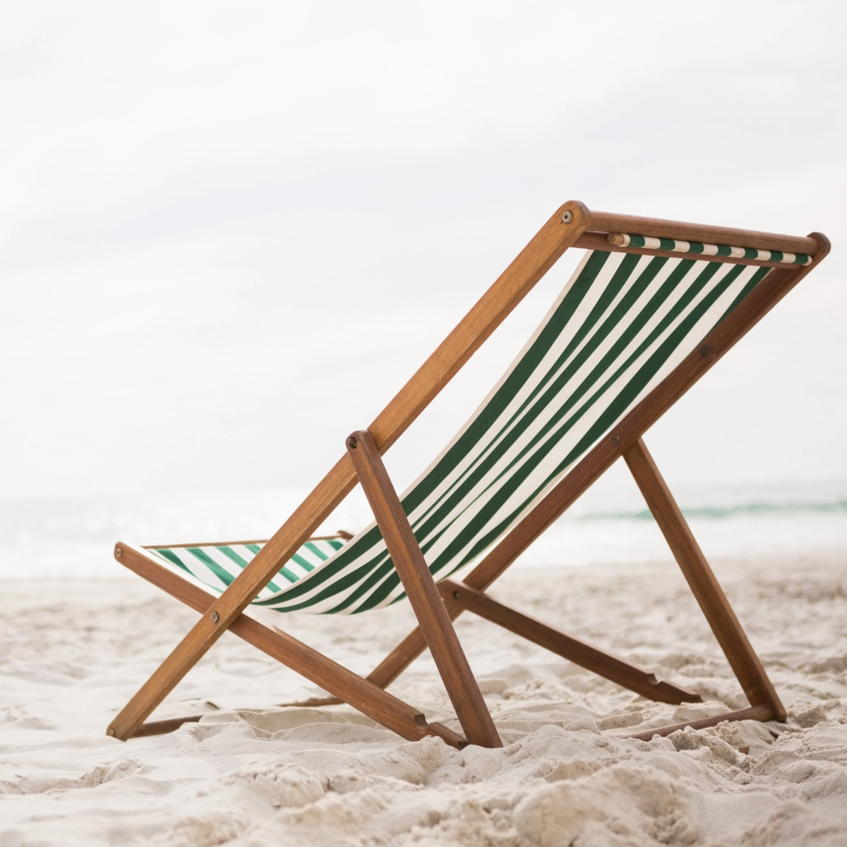 Classic wooden low beach chair with green striped fabric on a white sandy beach.