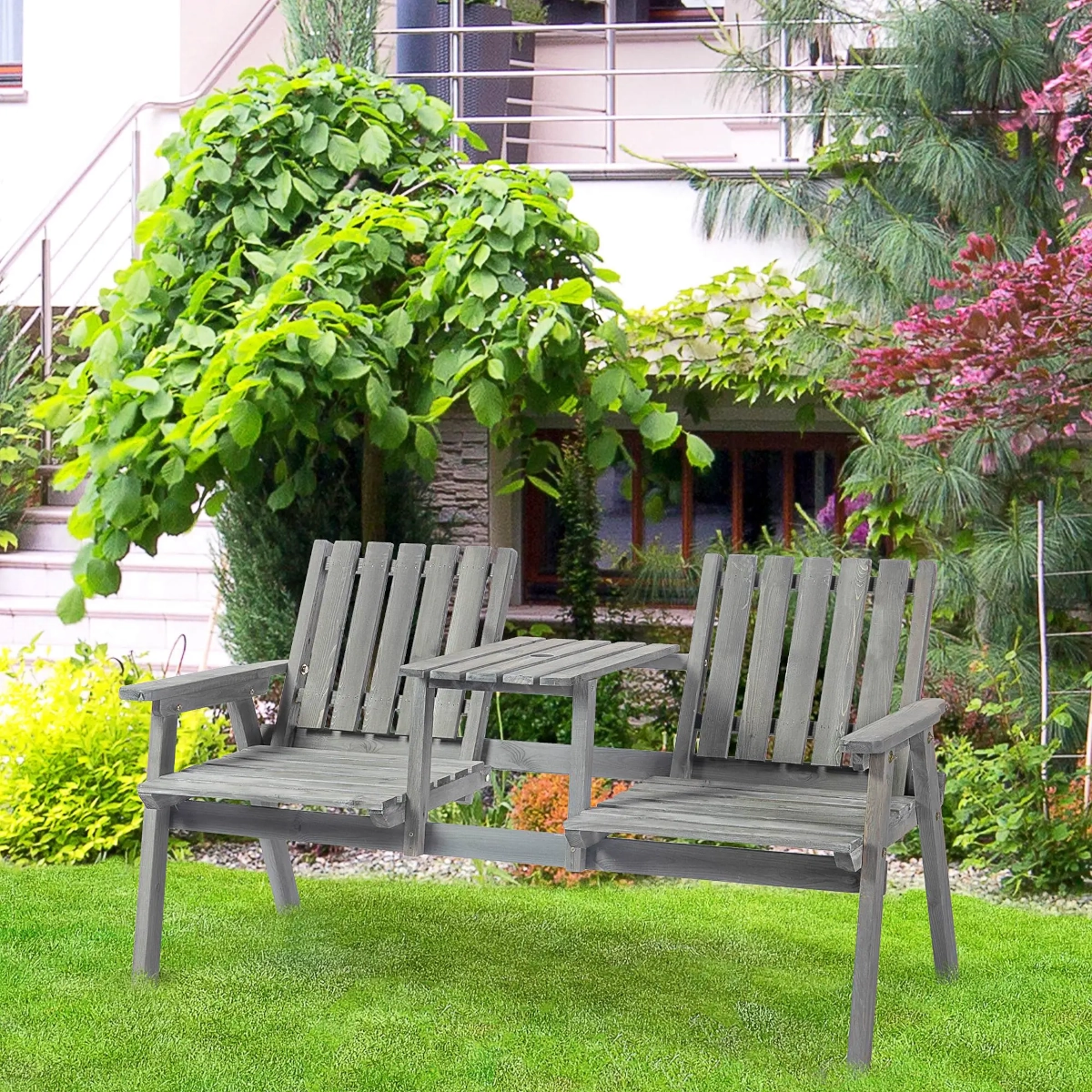 Grey two-seater wooden bench with central table placed on a lush green lawn.