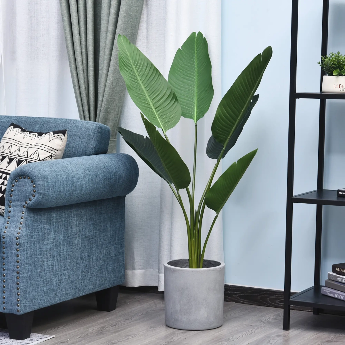 Large artificial bird of paradise plant in grey pot next to a blue armchair in living room.