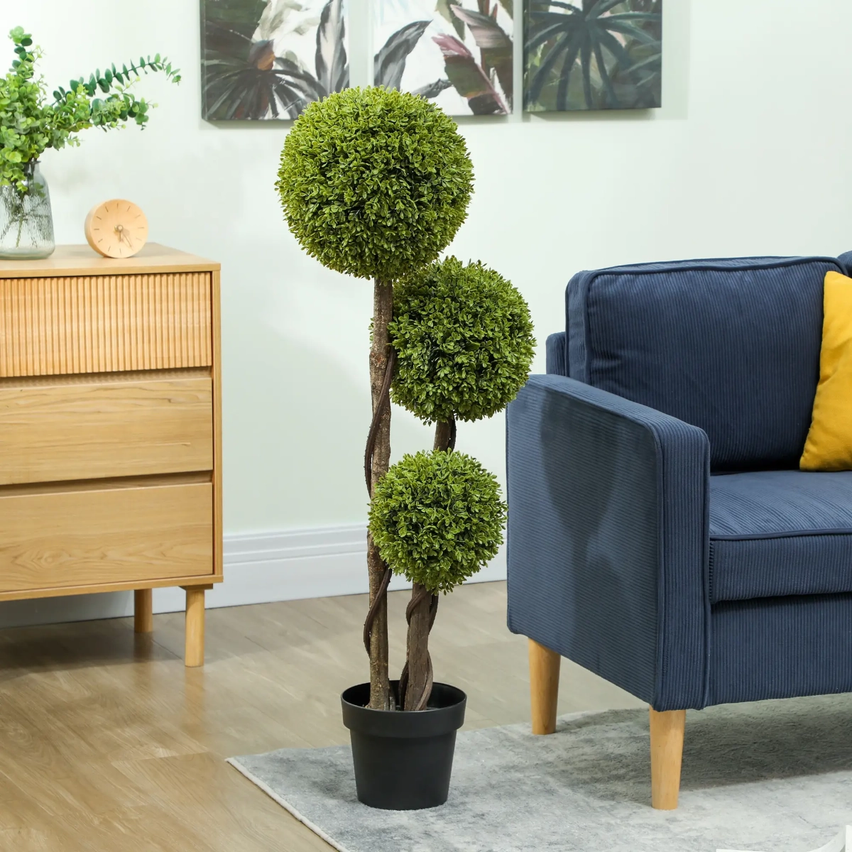 Three-ball artificial boxwood topiary tree in black pot beside blue armchair and wooden cabinet.