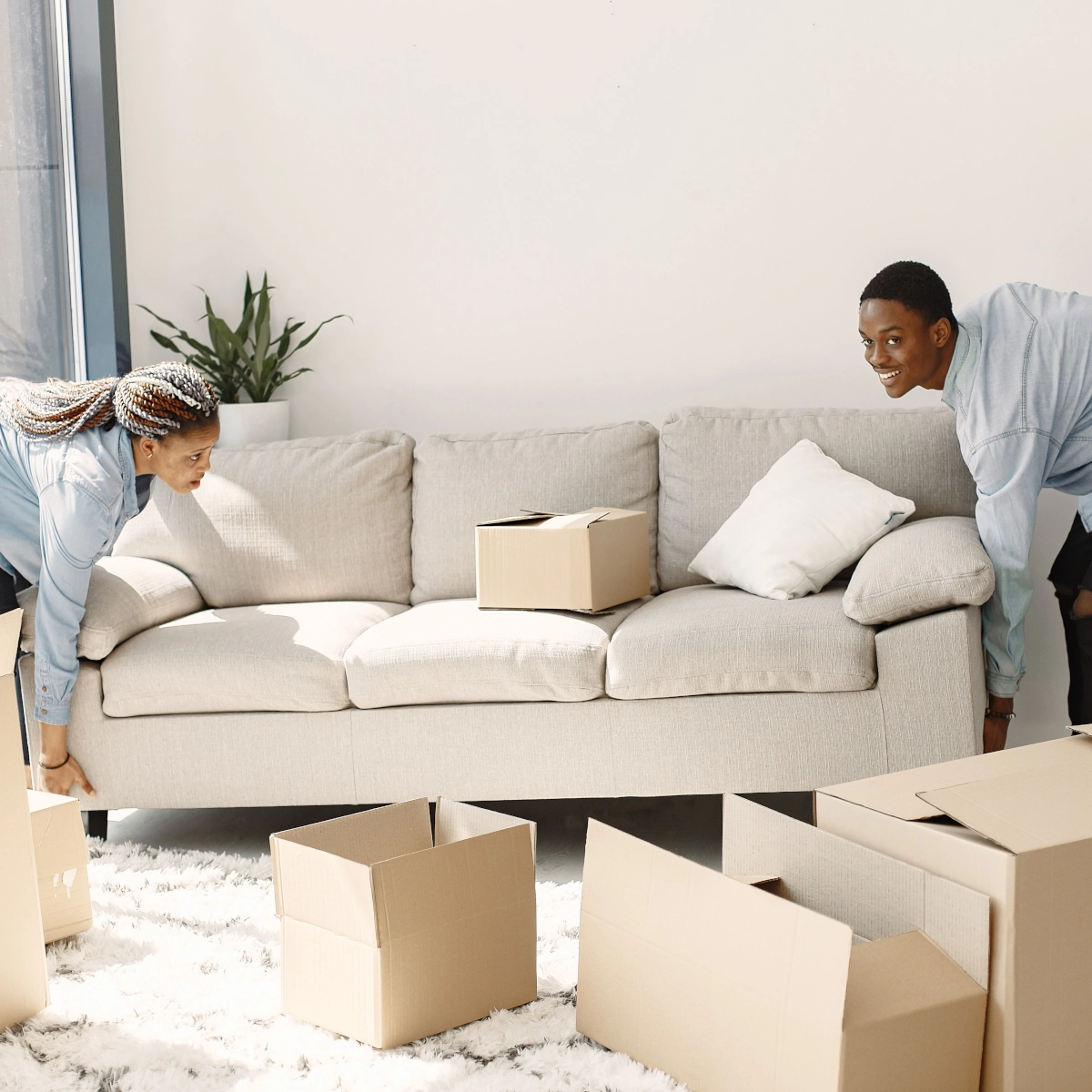 Two people moving a beige sofa surrounded by cardboard boxes in a bright room.