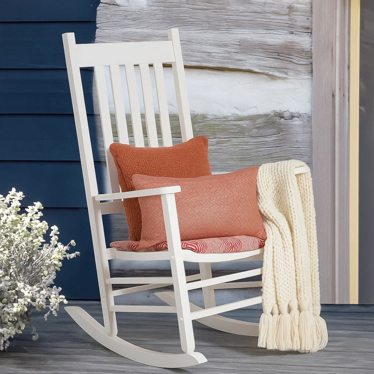 White rocking chair with orange cushions and a cream knit throw on a rustic wooden porch.