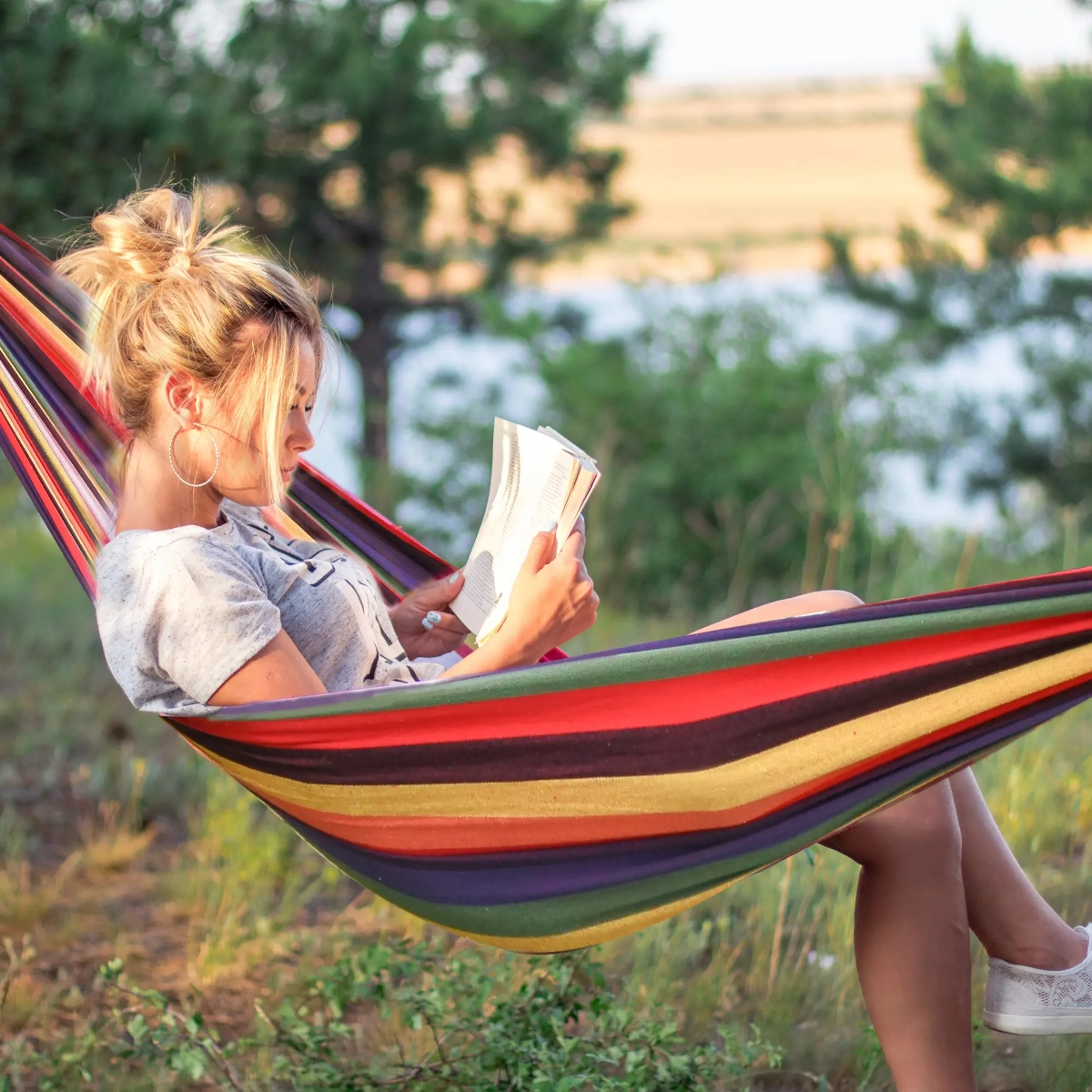 Woman relaxing in a striped hammock outdoors, reading a book by the lake.