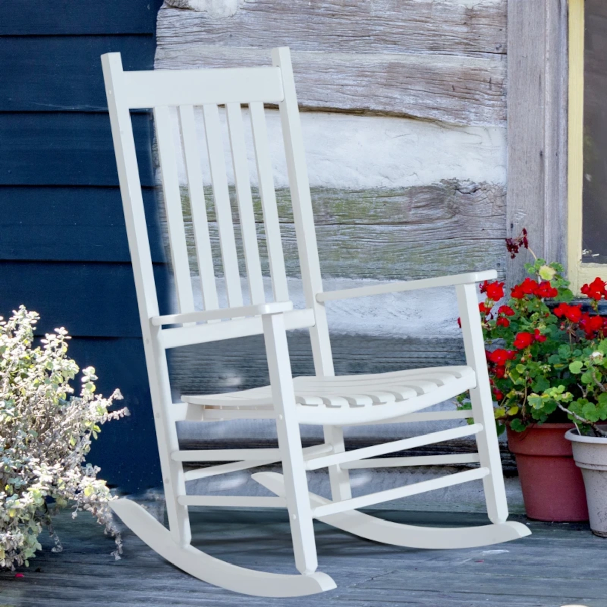 Simple white rocking chair on a wooden porch near flower pots and a weathered cabin wall.