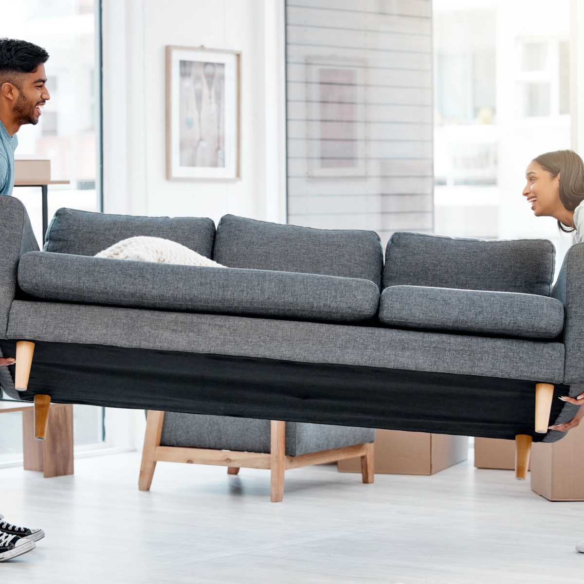 Two people lifting and moving a grey sofa in a bright living room.