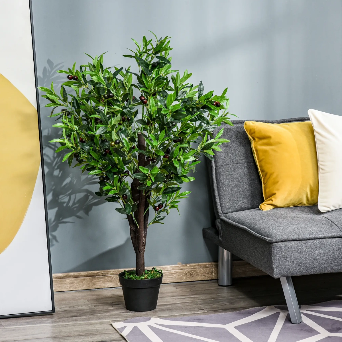  Lush artificial olive tree in black pot next to grey sofa with yellow and white pillows.