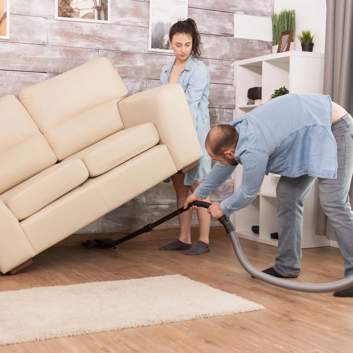Man vacuums floor under cream sofa as woman holds furniture, cleaning living room together