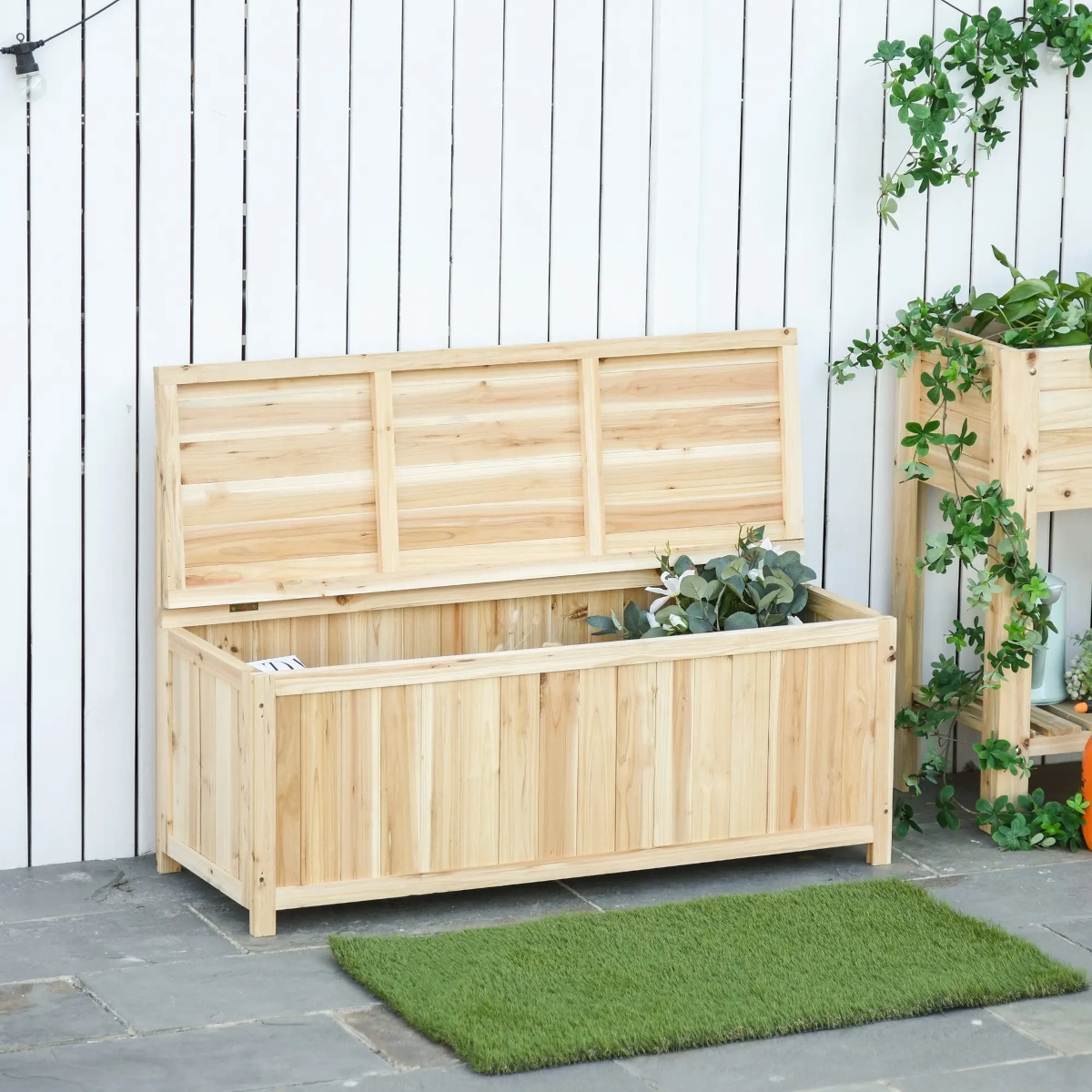 Natural wooden storage bench with open lid placed against a white fence in a garden.