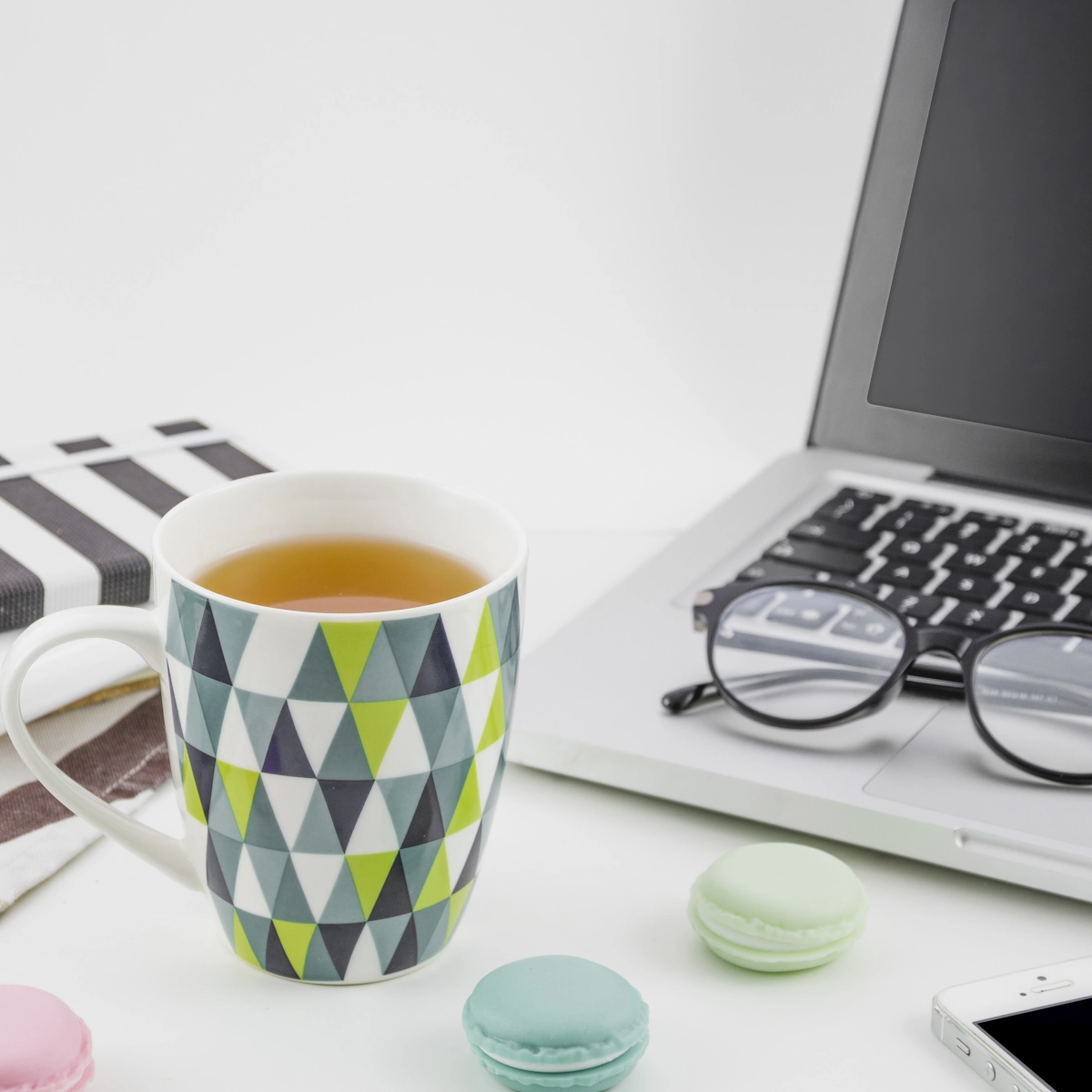 Patterned mug with tea near laptop, macarons, notebook, and eyeglasses