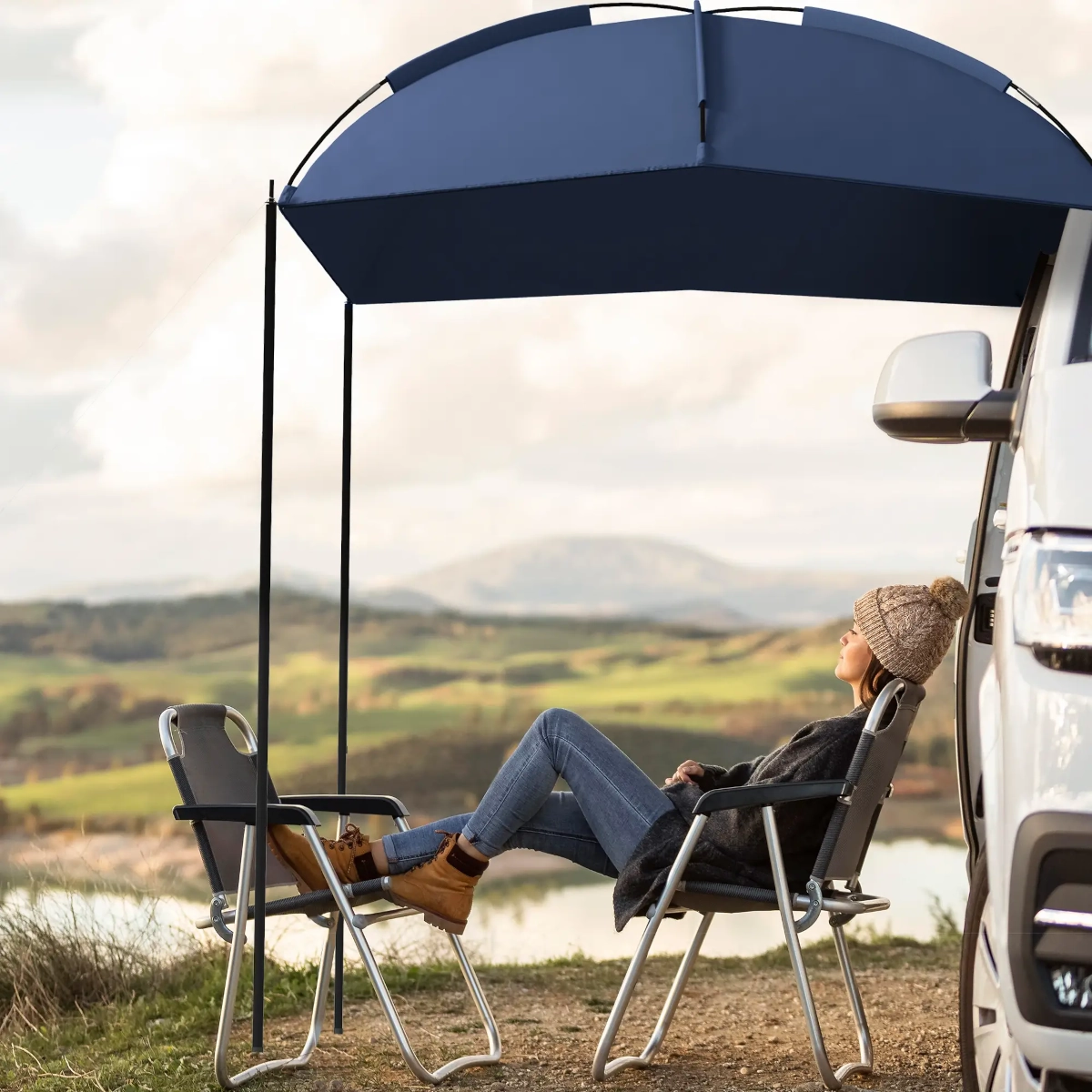 Woman relaxing in camping chair under vehicle awning with mountain and lake views.
