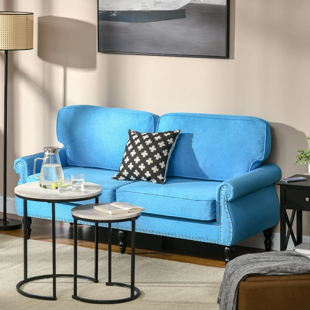 Blue two-seater sofa with studded arms and a patterned cushion beside modern nesting tables.