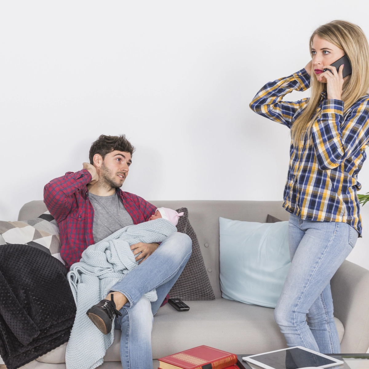 Man holding baby on couch looking tired, woman talking on phone in background.