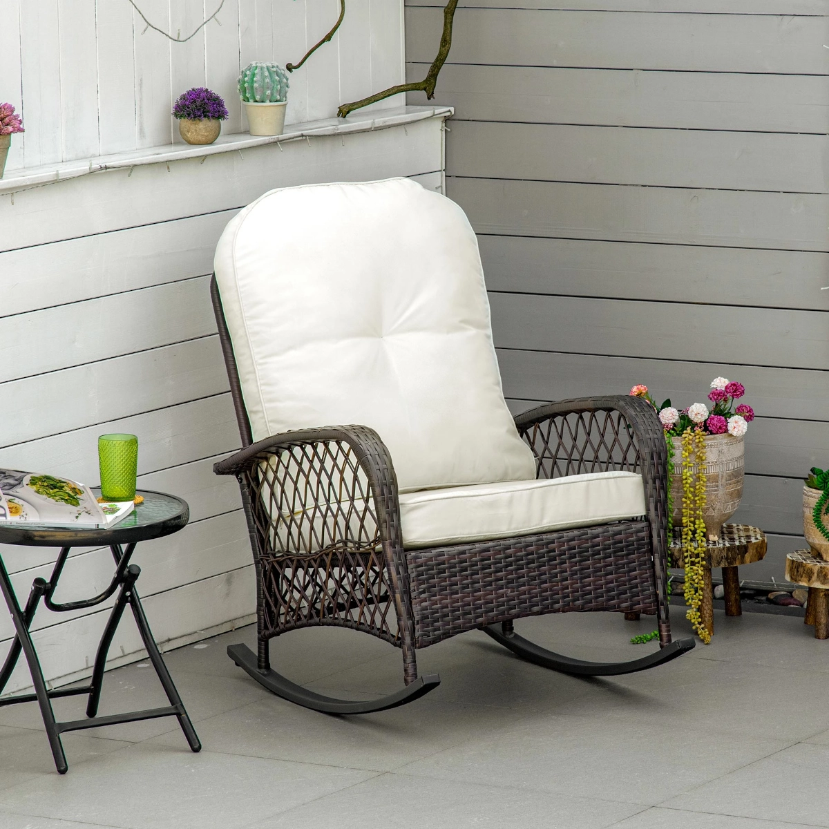 Stylish wicker rocking chair with white cushions on a tiled patio near decorative plants.