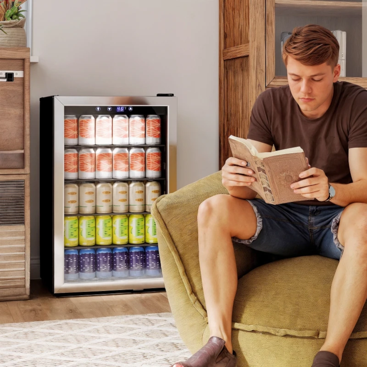 Man reading next to a fully stocked can beverage fridge in a cozy living space.