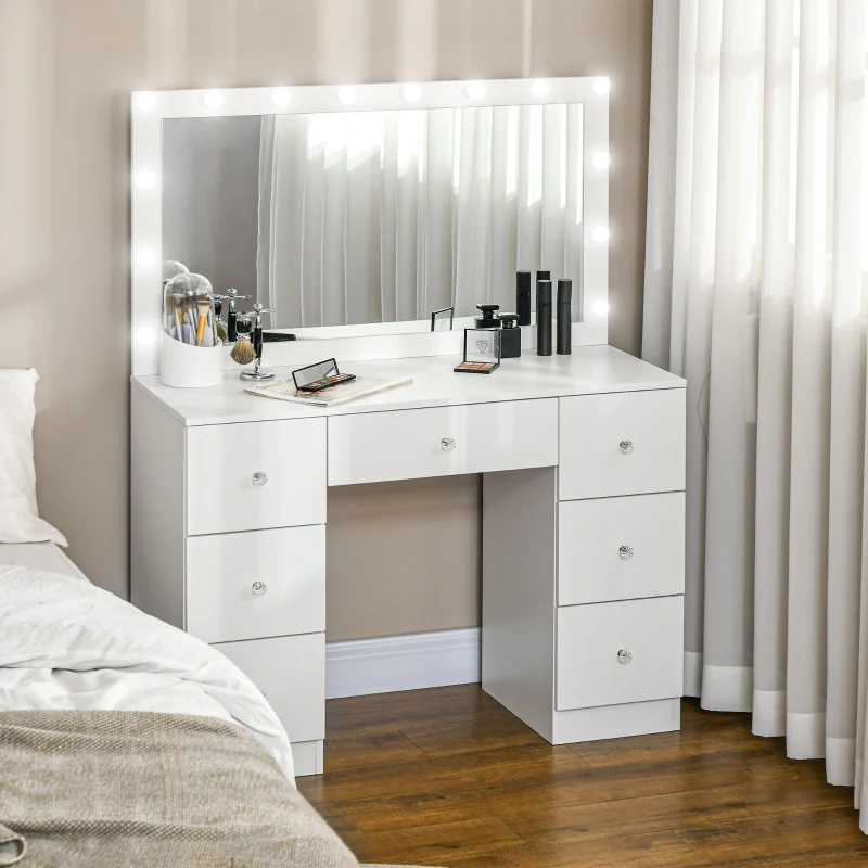 White vanity table with large lighted mirror, drawers, and makeup neatly displayed beside a bed.