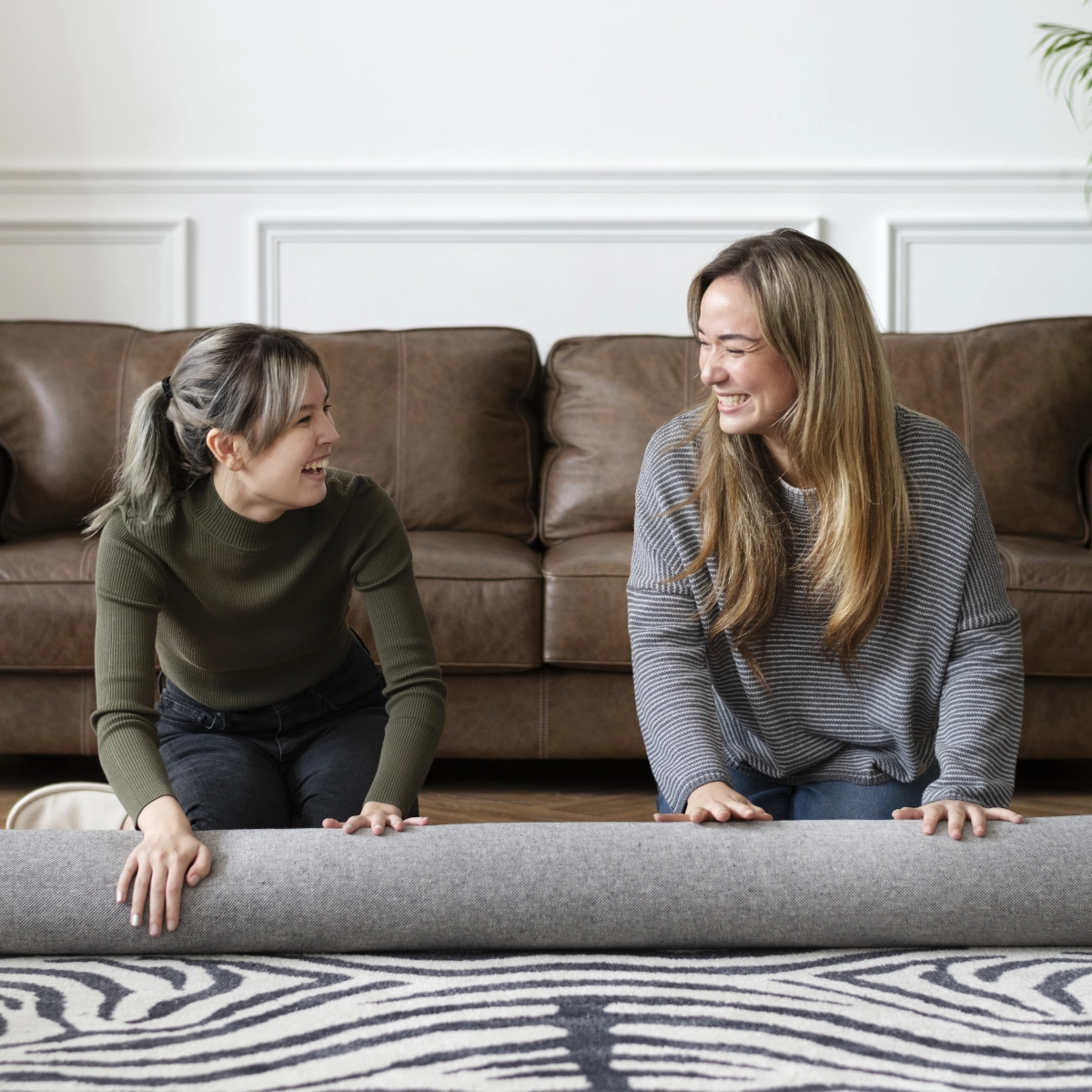 Two women working together to flatten an area rug on hardwood floor in a cozy living room.