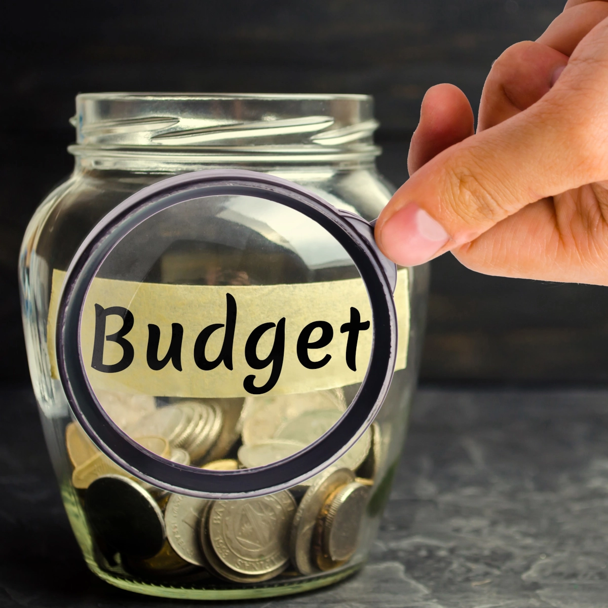 Glass jar labeled budget filled with coins and a hand holding magnifying glass in front.