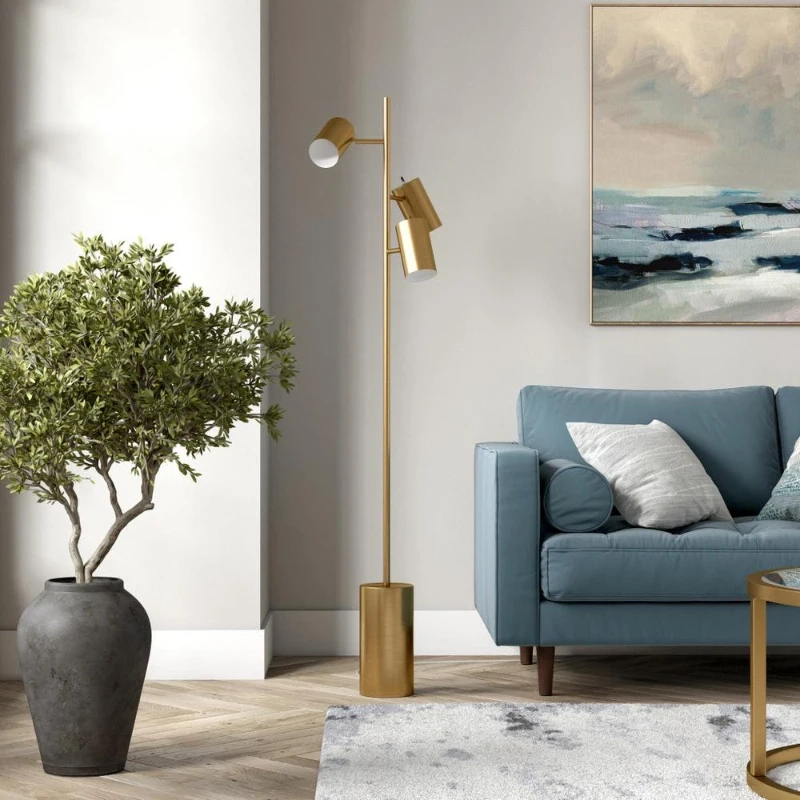 Modern living room with gold triple spotlight floor lamp beside blue sofa and large plant.