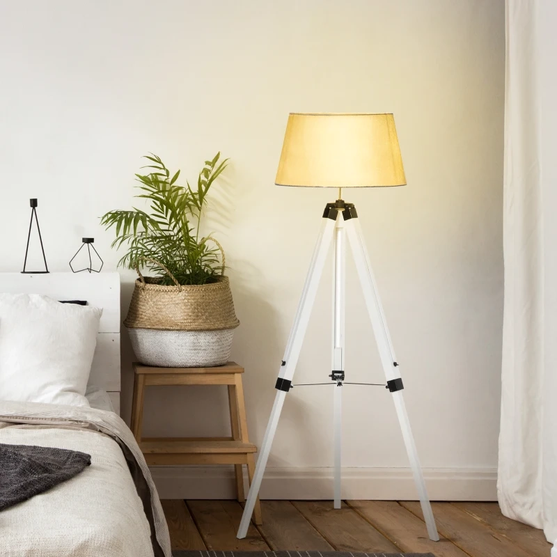 Cozy bedroom with white tripod floor lamp and fabric shade beside bed and potted plant.