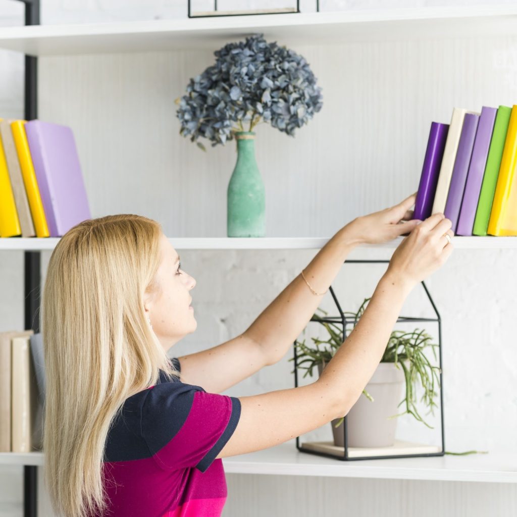 Woman organizing colorful books on modern white bookshelf with plants and décor.