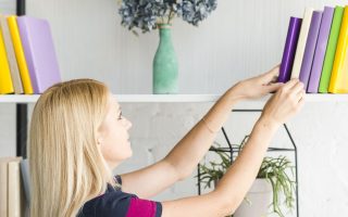 Woman organizing colorful books on modern white bookshelf with plants and décor.