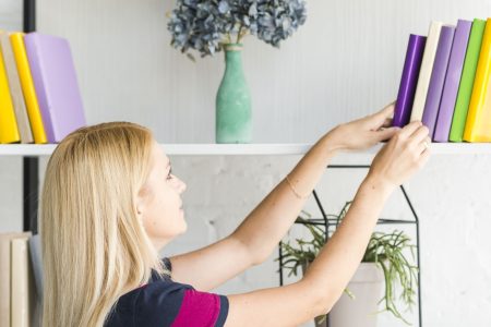 Woman organizing colorful books on modern white bookshelf with plants and décor.