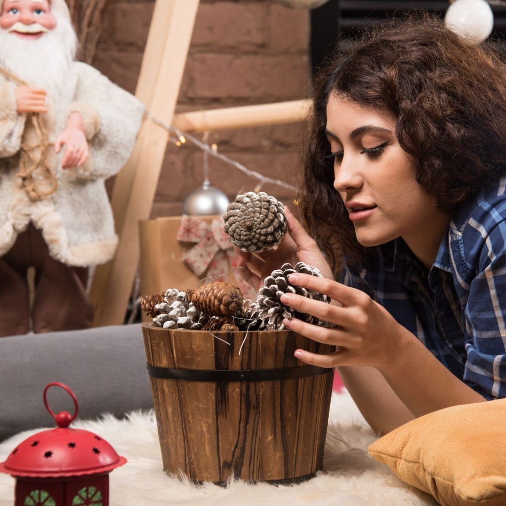 Woman arranging frosted pinecones in a wooden bucket for cozy holiday décor.