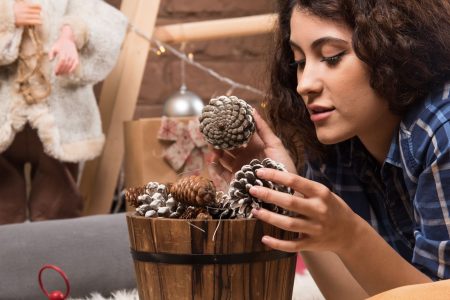 Woman arranging frosted pinecones in a wooden bucket for cozy holiday décor.