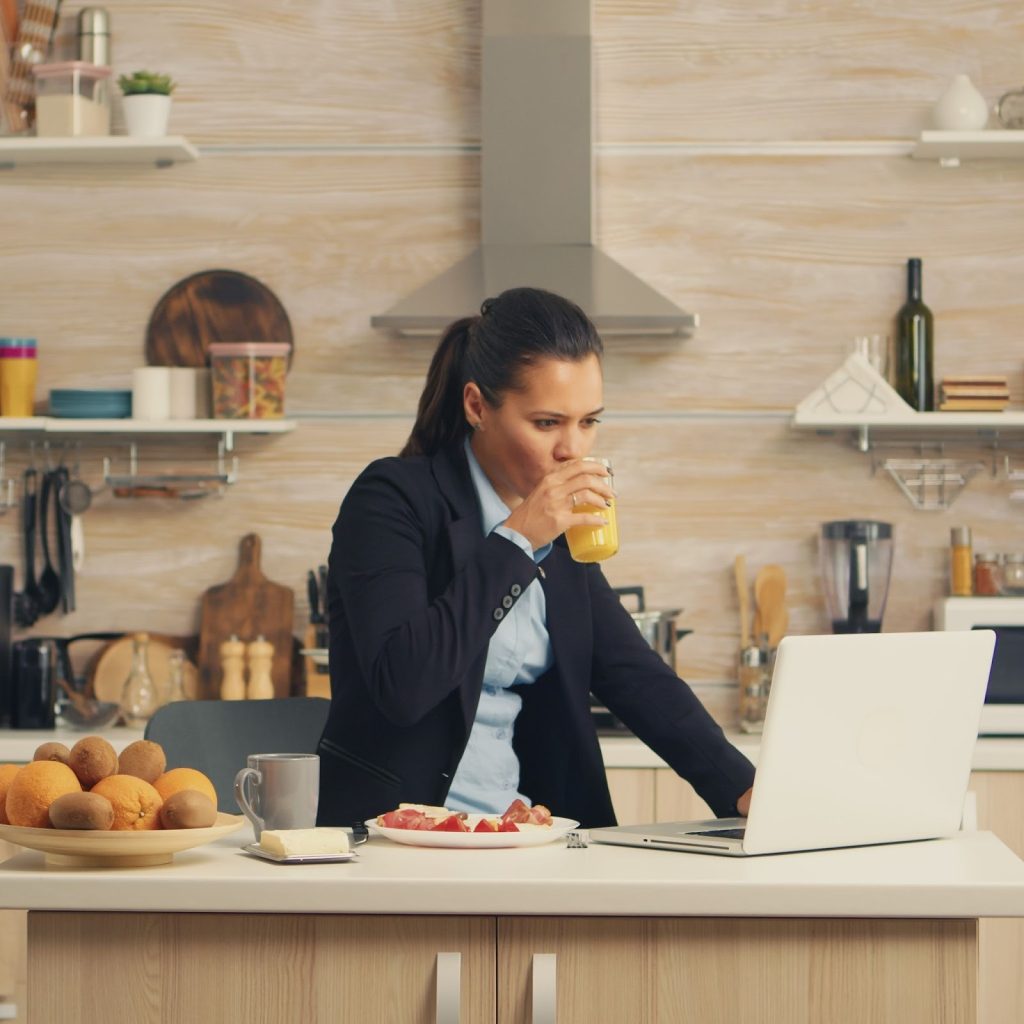 Woman working on laptop at kitchen island while having breakfast and multitasking.