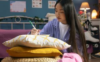 Student writing in colorful dorm room with cushions and décor.