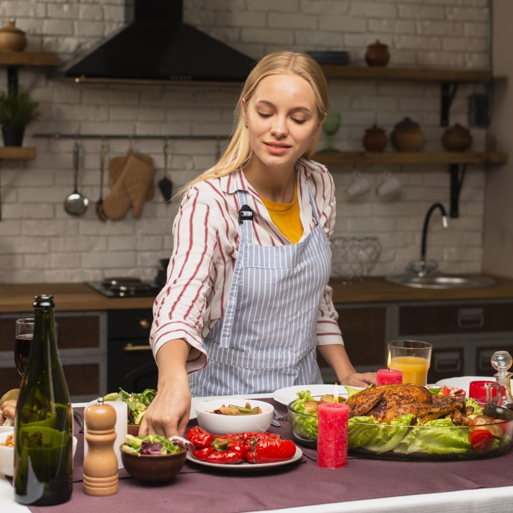 Woman preparing a festive buffet on a kitchen island with roasted vegetables, chicken, and table décor.